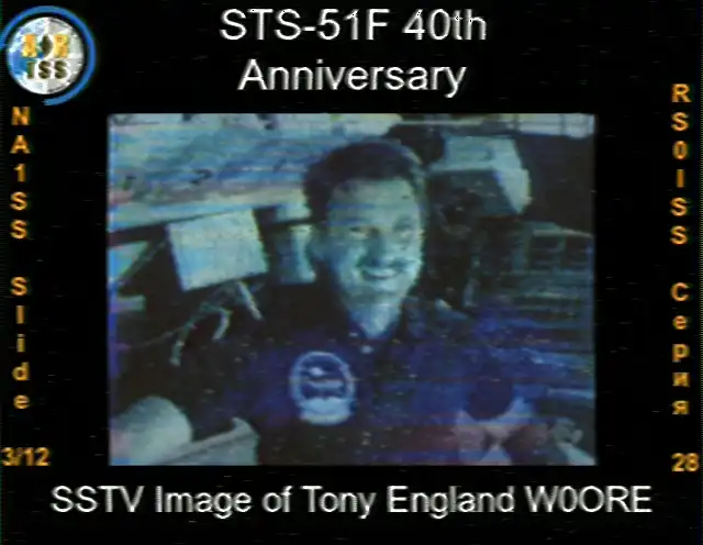 Obraz SSTV z ISS, slajd 3 z 12, 40. rocznica. Obraz przedstawia portret astronauty Tony'ego Englanda, który uśmiecha się i patrzy w kierunku aparatu. Ma na sobie niebieską koszulkę polo z naszywką misji na piersi. Tło ukazuje wnętrze promu. Tekst na dole identyfikuje obraz jako 'Obraz SSTV Tony'ego Englanda W0ORE'. Obraz ma lekko niebieskawy odcień.