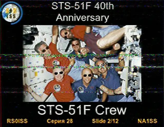 Obraz SSTV z ISS, slajd 2 z 12. Obraz przedstawia siedmioosobową załogę misji promu kosmicznego Challenger, unoszącą się razem w środowisku mikrograwitacji modułu Spacelab. Wszyscy astronauci mają na sobie okulary przeciwsłoneczne i uśmiechają się do grupowego zdjęcia. Tekst na dole głosi 'Załoga STS-51F'.