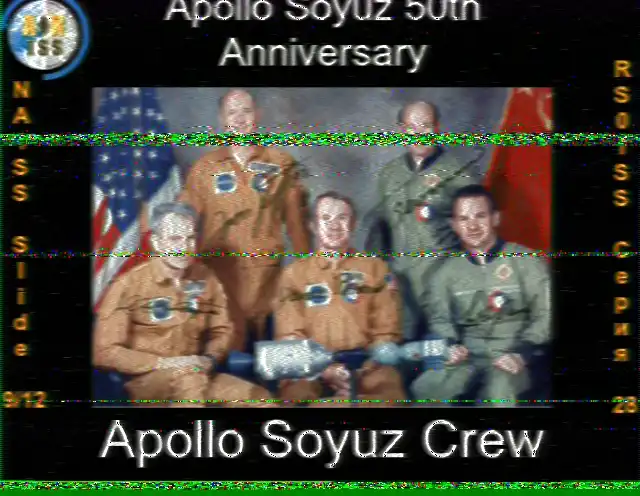 Obraz SSTV z ISS, slajd 9 z 12, upamiętniający 50. rocznicę misji Apollo-Sojuz. Obraz to formalny portret grupowy amerykańskich astronautów i radzieckich kosmonautów. Członkowie załogi pozują razem przed flagami amerykańską i radziecką, trzymając model zadokowanych statków Apollo i Sojuz. Tekst na dole głosi 'Załoga Apollo Sojuz'.